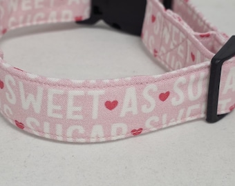 May include: A pink dog collar with the words "SWEET AS SUGAR" in white, interspersed with red heart and lip graphics. The collar has a black plastic buckle.
