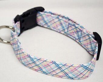 May include: A white dog collar with a blue, pink, and yellow plaid pattern. The collar has a black plastic buckle and a silver D-ring.
