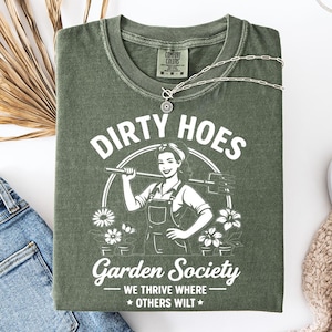 May include: An olive green t-shirt with the text "DIRTY HOES Garden Society" and a graphic of a woman with a hoe. The shirt is folded with a silver necklace, blue jeans, and white sneakers. A dried palm leaf is also in the image.