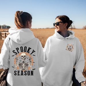 May include: White hoodie with a Halloween-themed design. The back features a skeleton with peace signs, the words "Spooky Season," and bats. The front has a smaller version of the design. The image is taken outdoors with a field in the background.