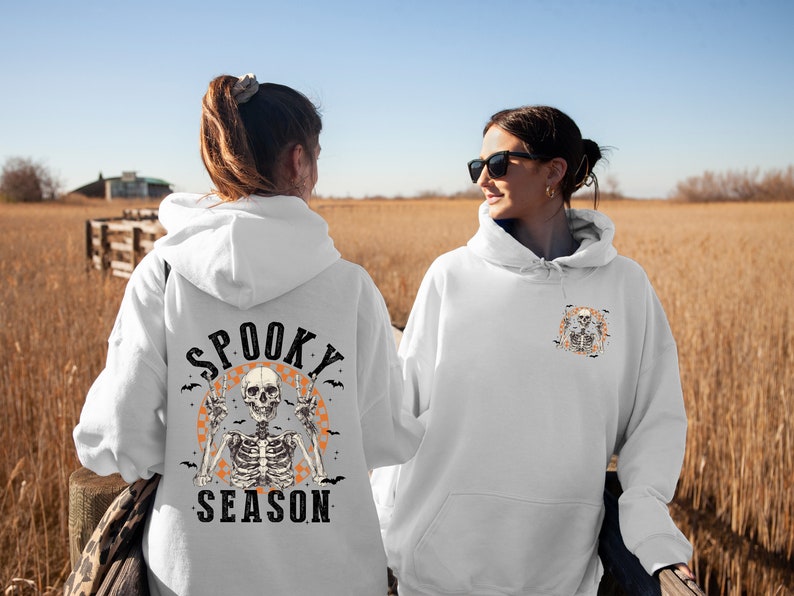 May include: White hoodie with a Halloween-themed design. The back features a skeleton with peace signs, the words "Spooky Season," and bats. The front has a smaller version of the design. The image is taken outdoors with a field in the background.