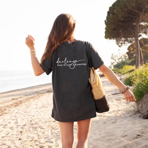 May include: A woman wearing a dark gray t-shirt with the text "darling this is just a chapter" printed on the back. She is walking on a sandy beach with a woven tote bag.