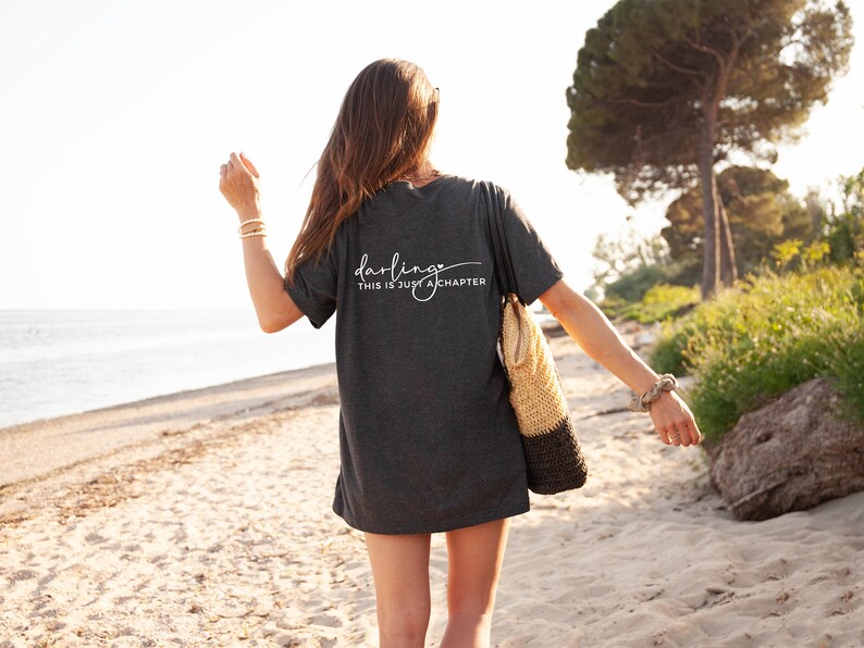 May include: A woman wearing a dark gray t-shirt with the text "darling this is just a chapter" printed on the back. She is walking on a sandy beach with a woven tote bag.