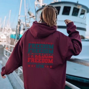 May include: A maroon hoodie with a large graphic print on the back. The graphic features the text "Land of the Free" and "Freedom" repeated four times in a vintage style font. The text is in red and blue with a white star pattern. The text "Because of the Brave" and "1776" are also included in the graphic.