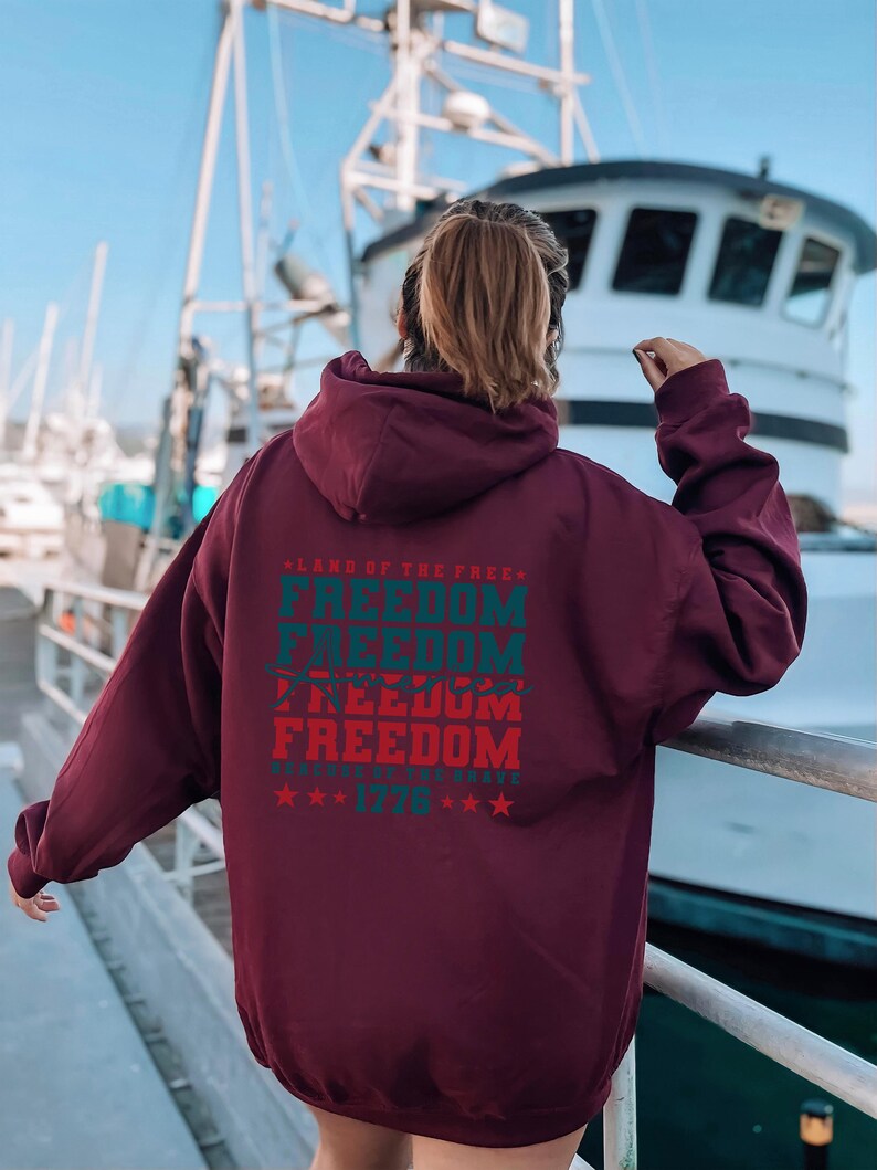 May include: A maroon hoodie with a large graphic print on the back. The graphic features the text "Land of the Free" and "Freedom" repeated four times in a vintage style font. The text is in red and blue with a white star pattern. The text "Because of the Brave" and "1776" are also included in the graphic.