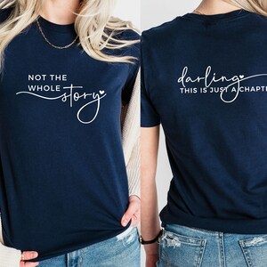 May include: A navy blue t-shirt with the text "Not the whole story" on the front and "Darling, this is just a chapter" on the back. Both phrases are written in a cursive font with a small heart symbol.