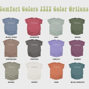 May include: A display of Comfort Colors 1717 t-shirts in various colors, including blue jean, crimson, bay, moss, espresso, pepper, violet, berry, khaki, ivory, yam, and blue spruce. Each shirt has the text "love like Jesus".
