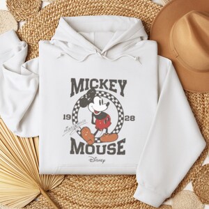 May include: White hooded sweatshirt with a vintage Mickey Mouse graphic. The graphic features Mickey Mouse in a red and black outfit, standing in a circle with the text "Mickey Mouse" and "1928" printed around him.