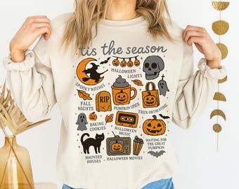 May include: Beige sweatshirt with a Halloween-themed graphic. The design includes a witch on a crescent moon, a skull, pumpkins, and the phrase "'tis the season." Other elements include a black cat, a tombstone, and the words "Spooky Witches" and "Haunted Houses."