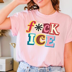 May include: Pink t-shirt with the words "f*ck ice" in colorful, retro-style lettering. The text features a mix of fonts and colors, including yellow, red, black, and blue. The shirt is made of a soft material.
