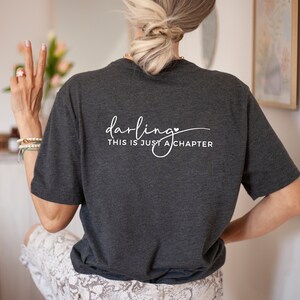 May include: A dark gray t-shirt with the text "darling this is just a chapter" in white lettering with a small heart symbol.