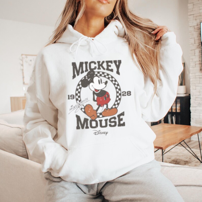 May include: White hooded sweatshirt with a vintage Mickey Mouse graphic. The graphic features Mickey Mouse in a red and black outfit with a black and white checkered circle around him. The text "Mickey Mouse" is printed below the graphic with the years "1928" and "1928" on either side of the circle. The text "Disney" is printed below the text "Mickey Mouse".