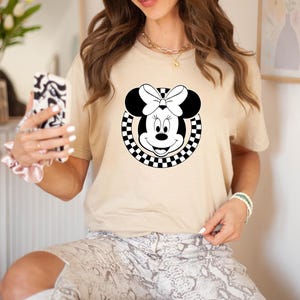 May include: A beige t-shirt with a black and white checkered circle design featuring a cartoon image of Minnie Mouse with a black bow.