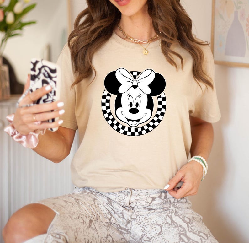 May include: A beige t-shirt with a black and white checkered circle design featuring a cartoon image of Minnie Mouse with a black bow.