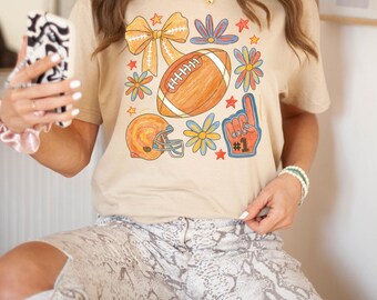 May include: Beige t-shirt featuring a football, helmet, bow, flowers, and a foam finger with the number one. The graphic is in shades of orange, blue, and yellow. The shirt is paired with snake print pants.