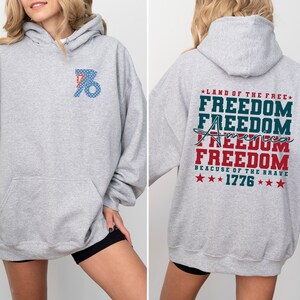 May include: A grey hooded sweatshirt with a red, white and blue graphic on the back that says "Land of the Free" and "Freedom" four times, with "America" written in cursive and "Because of the Brave" and "1776" below. The front of the sweatshirt has a red, white and blue graphic of the number "76" with stars.
