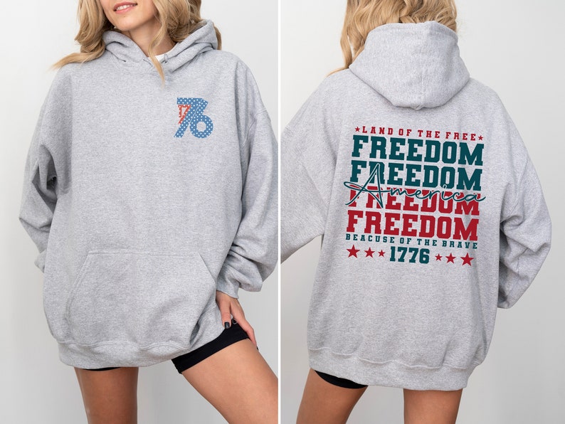 May include: A grey hooded sweatshirt with a red, white and blue graphic on the back that says "Land of the Free" and "Freedom" four times, with "America" written in cursive and "Because of the Brave" and "1776" below. The front of the sweatshirt has a red, white and blue graphic of the number "76" with stars.
