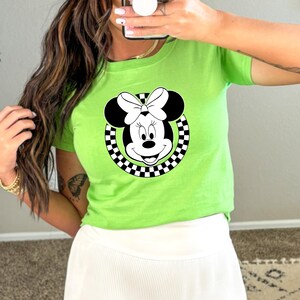 May include: A green t-shirt with a black and white checkered circle around a cartoon image of Minnie Mouse. Minnie Mouse is wearing a black bow.