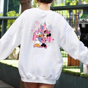 K&ouml;nnte beinhalten: Wei&szlig;es Sweatshirt mit einem farbenfrohen Aufdruck von Minnie Mouse und Daisy Duck vor einem Schloss. Die Grafik enth&auml;lt rosa, lila und gelbe Akzente. Das Sweatshirt hat lange &Auml;rmel und einen Rundhalsausschnitt.