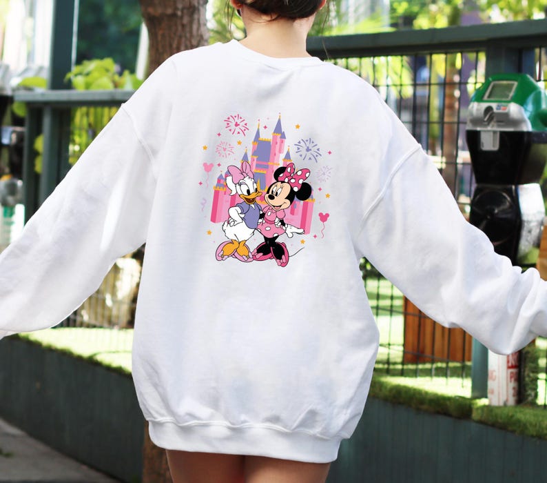 K&ouml;nnte beinhalten: Wei&szlig;es Sweatshirt mit einem farbenfrohen Aufdruck von Minnie Mouse und Daisy Duck vor einem Schloss. Die Grafik enth&auml;lt rosa, lila und gelbe Akzente. Das Sweatshirt hat lange &Auml;rmel und einen Rundhalsausschnitt.