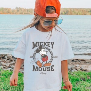 May include: White t-shirt with a vintage Mickey Mouse graphic. The graphic features Mickey Mouse in a red and black outfit, standing in a circle with the text "Mickey Mouse" and "1928" and "Disney".