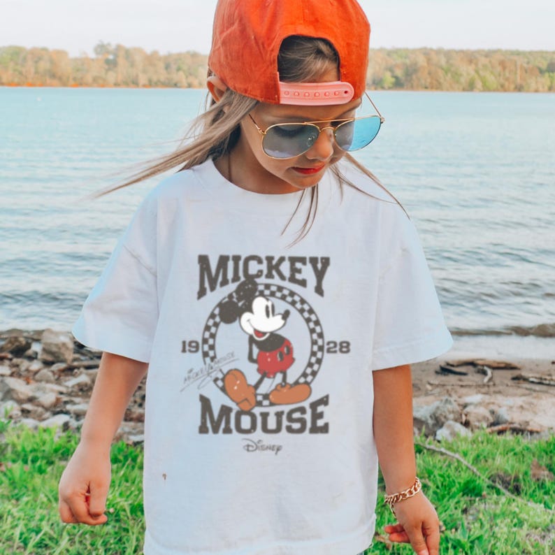 May include: White t-shirt with a vintage Mickey Mouse graphic. The graphic features Mickey Mouse in a red and black outfit, standing in a circle with the text "Mickey Mouse" and "1928" and "Disney".