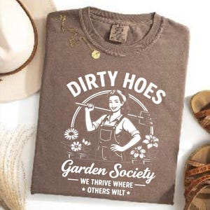 May include: Brown t-shirt with the text "DIRTY HOES" above a graphic of a woman holding a gardening tool. The text "Garden Society" and "WE THRIVE WHERE * OTHERS WILT *" are below the graphic. A gold necklace and a hat are visible.