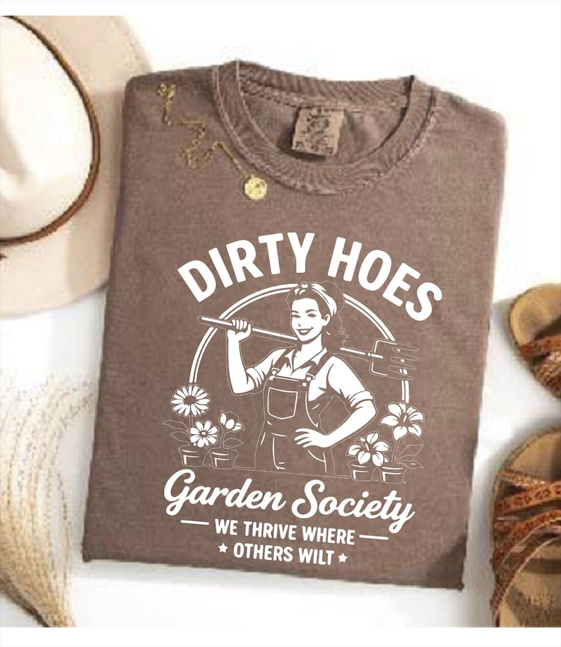 May include: Brown t-shirt with the text "DIRTY HOES" above a graphic of a woman holding a gardening tool. The text "Garden Society" and "WE THRIVE WHERE * OTHERS WILT *" are below the graphic. A gold necklace and a hat are visible.