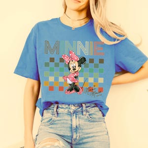 Puede incluir: Camiseta azul con un dise&ntilde;o de Minnie Mouse. El dise&ntilde;o incluye la palabra "MINNIE" en una fuente colorida, un patr&oacute;n de cuadros y una ilustraci&oacute;n de Minnie Mouse. La camiseta tambi&eacute;n tiene la firma "Minnie Mouse xoxo".