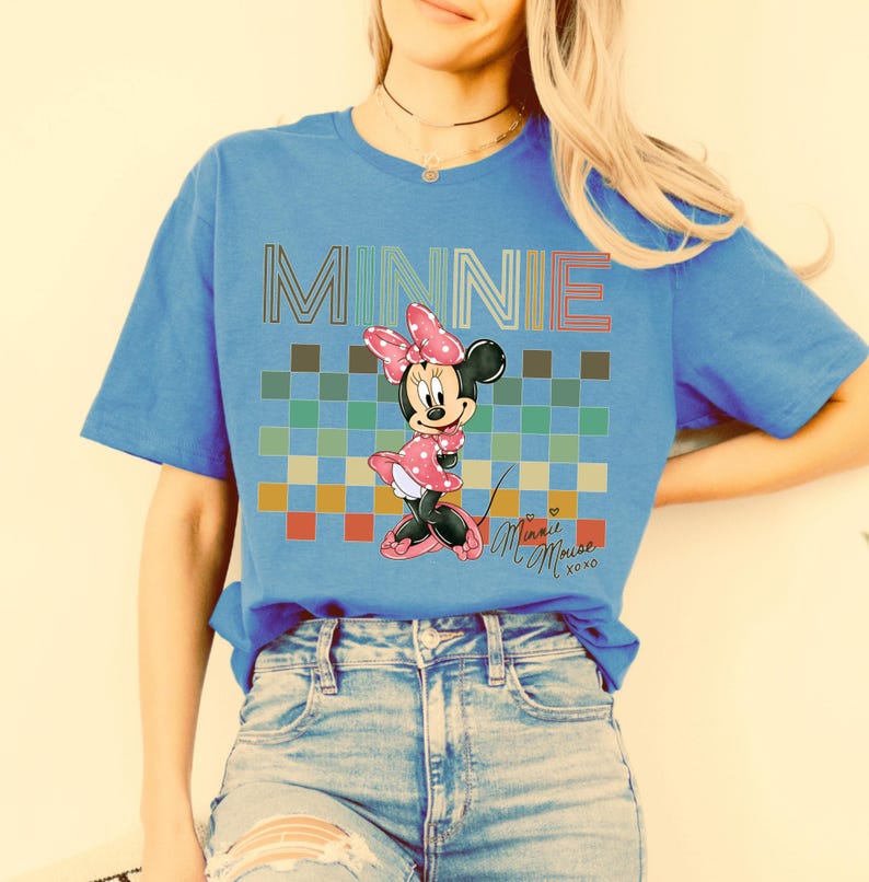 Puede incluir: Camiseta azul con un dise&ntilde;o de Minnie Mouse. El dise&ntilde;o incluye la palabra "MINNIE" en una fuente colorida, un patr&oacute;n de cuadros y una ilustraci&oacute;n de Minnie Mouse. La camiseta tambi&eacute;n tiene la firma "Minnie Mouse xoxo".