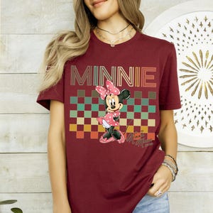 Puede incluir: Camiseta burdeos con la palabra "MINNIE" en una fuente retro sobre un patr&oacute;n de cuadros. La camiseta muestra un personaje de dibujos animados de Minnie Mouse con un vestido rosa de lunares. La camiseta tiene mangas cortas.