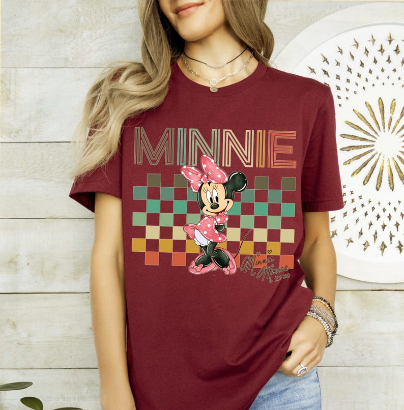 Puede incluir: Camiseta burdeos con la palabra "MINNIE" en una fuente retro sobre un patr&oacute;n de cuadros. La camiseta muestra un personaje de dibujos animados de Minnie Mouse con un vestido rosa de lunares. La camiseta tiene mangas cortas.