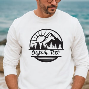 May include: White sweatshirt with a black mountain scene graphic and the text "Custom Text" in a circle.