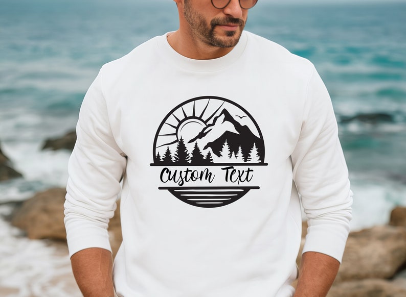 May include: White sweatshirt with a black mountain scene graphic and the text "Custom Text" in a circle.