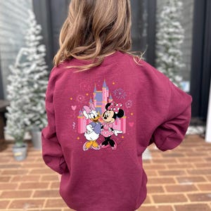 K&ouml;nnte beinhalten: Burgunderfarbenes Sweatshirt mit einem farbenfrohen Aufdruck von Daisy Duck und Minnie Mouse vor einem Schloss, mit Feuerwerk und Herzen. Das Sweatshirt ist aus weichem Material.