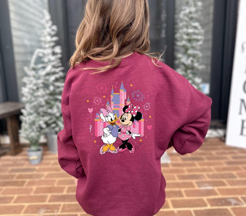 K&ouml;nnte beinhalten: Burgunderfarbenes Sweatshirt mit einem farbenfrohen Aufdruck von Daisy Duck und Minnie Mouse vor einem Schloss, mit Feuerwerk und Herzen. Das Sweatshirt ist aus weichem Material.