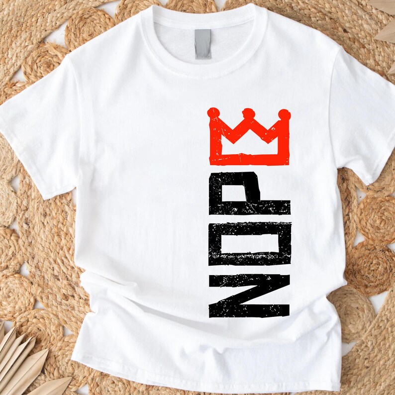 May include: White t-shirt with a red crown graphic above the black letters "NDR". The t-shirt is made of a soft material and has a crew neck. The design is printed vertically on the left side of the shirt.
