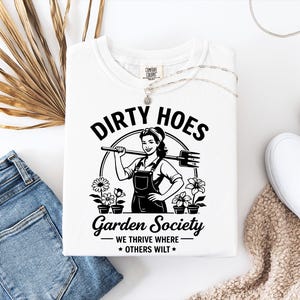 May include: White t-shirt with the text "DIRTY HOES Garden Society" and a graphic of a woman holding a garden tool. The shirt also features the phrase "WE THRIVE WHERE OTHERS WILT".