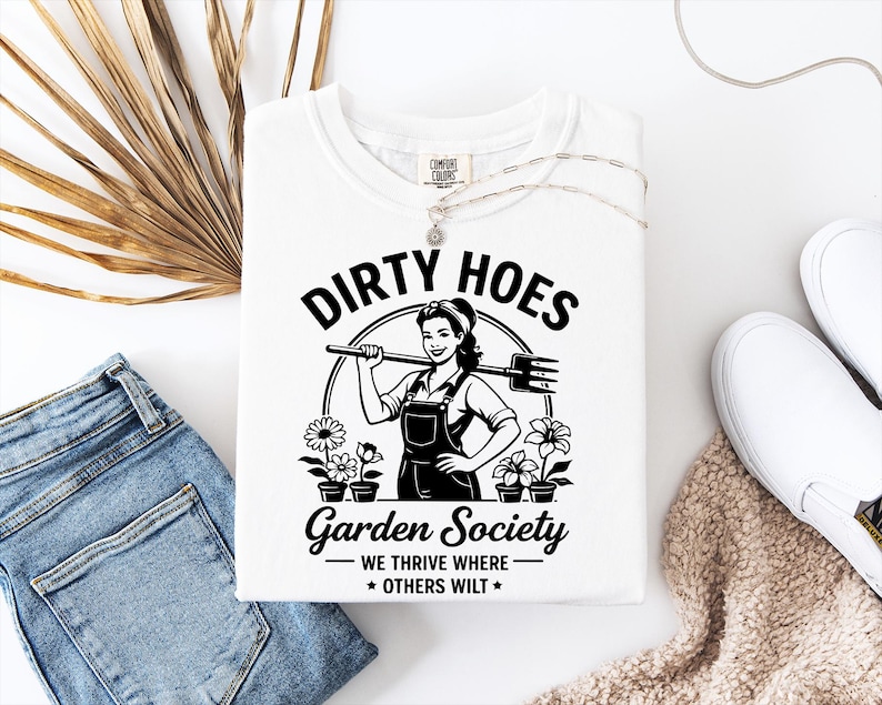 May include: White t-shirt with the text "DIRTY HOES Garden Society" and a graphic of a woman holding a garden tool. The shirt also features the phrase "WE THRIVE WHERE OTHERS WILT".