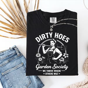 May include: Black t-shirt with the words "Dirty Hoes Garden Society" in white, featuring a graphic of a woman holding a gardening tool. The shirt also has the phrase "We thrive where others wilt."