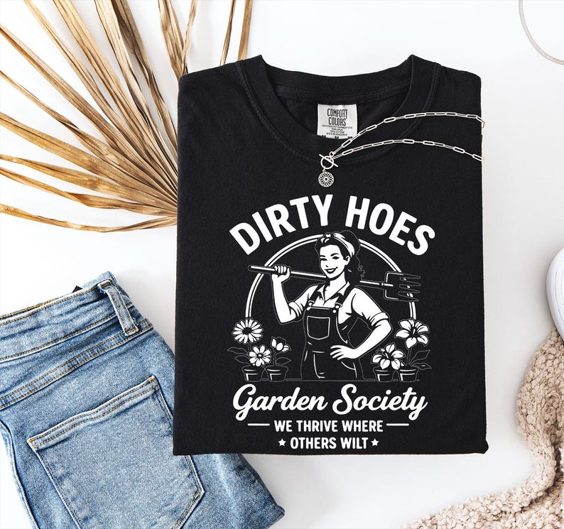 May include: Black t-shirt with the words "Dirty Hoes Garden Society" in white, featuring a graphic of a woman holding a gardening tool. The shirt also has the phrase "We thrive where others wilt."