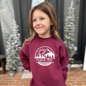 May include: A maroon sweatshirt with a white graphic of a mountain range, sun, and trees. The text "Custom Text" is printed below the graphic.