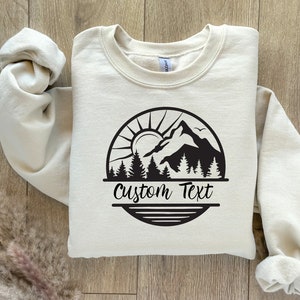 May include: A beige sweatshirt with a black mountain scene graphic and the text "Custom Text" printed on the front.