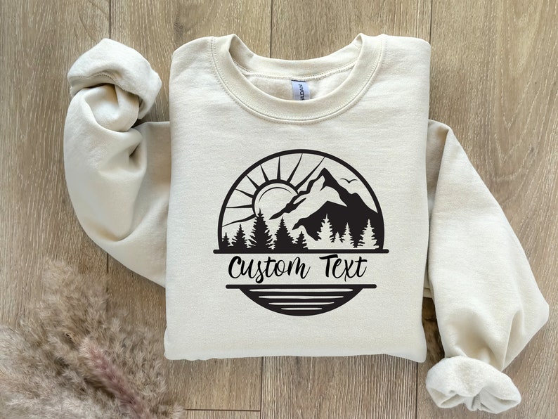 May include: A beige sweatshirt with a black mountain scene graphic and the text "Custom Text" printed on the front.