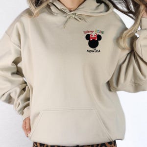 May include: A beige hoodie with a black and red Mickey Mouse graphic and the text "Disney 2025" above the graphic and "Monica" below the graphic.