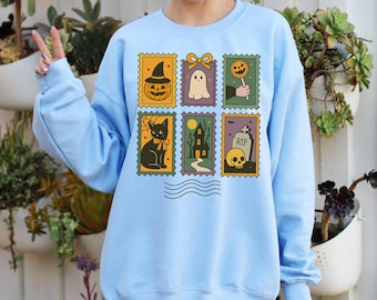 May include: Light blue sweatshirt featuring six Halloween-themed postage stamp designs. The stamps depict a jack-o'-lantern, ghost, lollipop, black cat, haunted house, and skull with a tombstone. The sweatshirt has long sleeves and a crew neck.