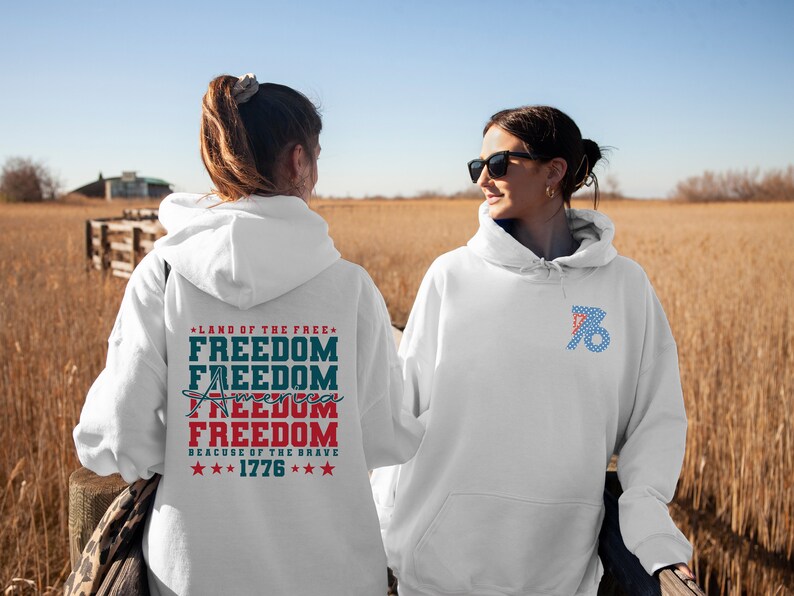 May include: Two white hooded sweatshirts with patriotic designs. One sweatshirt has the text "Land of the Free, Freedom, Freedom, America, Freedom, Because of the Brave, 1776" printed on the back. The other sweatshirt has a blue and red design with stars on the front.