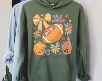 May include: Olive green hoodie with a football-themed graphic. The design includes a football, helmet, bow, flowers, and a foam finger. The graphic is in shades of orange, yellow, and blue.