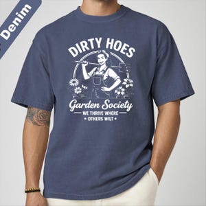 May include: Denim blue t-shirt with a white graphic that reads "Dirty Hoes Garden Society" featuring a woman with a gardening tool. The text "We thrive where others wilt" is also included in the design.
