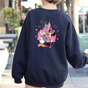 K&ouml;nnte beinhalten: Marineblaues Sweatshirt mit einem farbenfrohen Aufdruck von Daisy Duck und Minnie Mouse vor einem Schloss mit Feuerwerk. Die Grafik ist in Pink-, Lila- und Gelbt&ouml;nen gehalten. Das Sweatshirt hat lange &Auml;rmel.